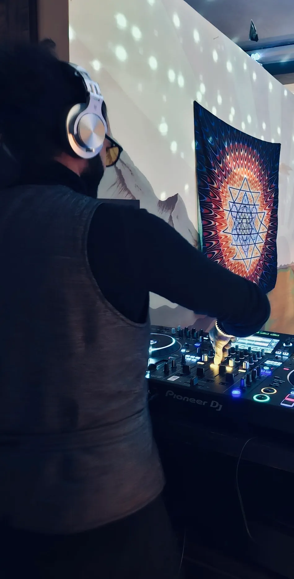 Sohrab Papahn mit Kopfhörern und Brille von hinten am DJ-Pult, an der Wand hängt ein buntes Tuch mit Sternmuster
