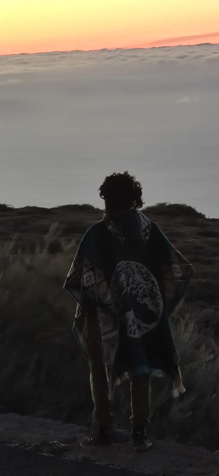 Sohrab Papahn mit Poncho von hinten auf einem Hügel, Blick auf Wolkendecke und Abendhimmel