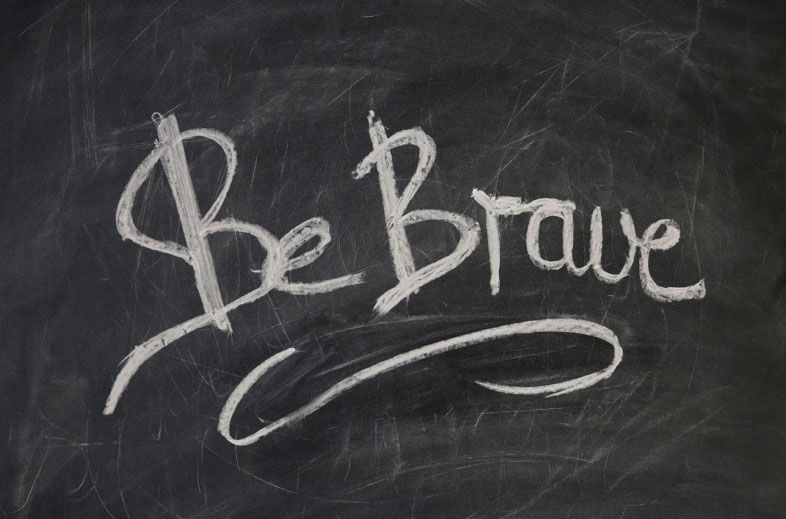 Der Schriftzug Be Brave in weißer Kreideschrift auf einer dunklen Tafel, darunter ein geschwungener Strich