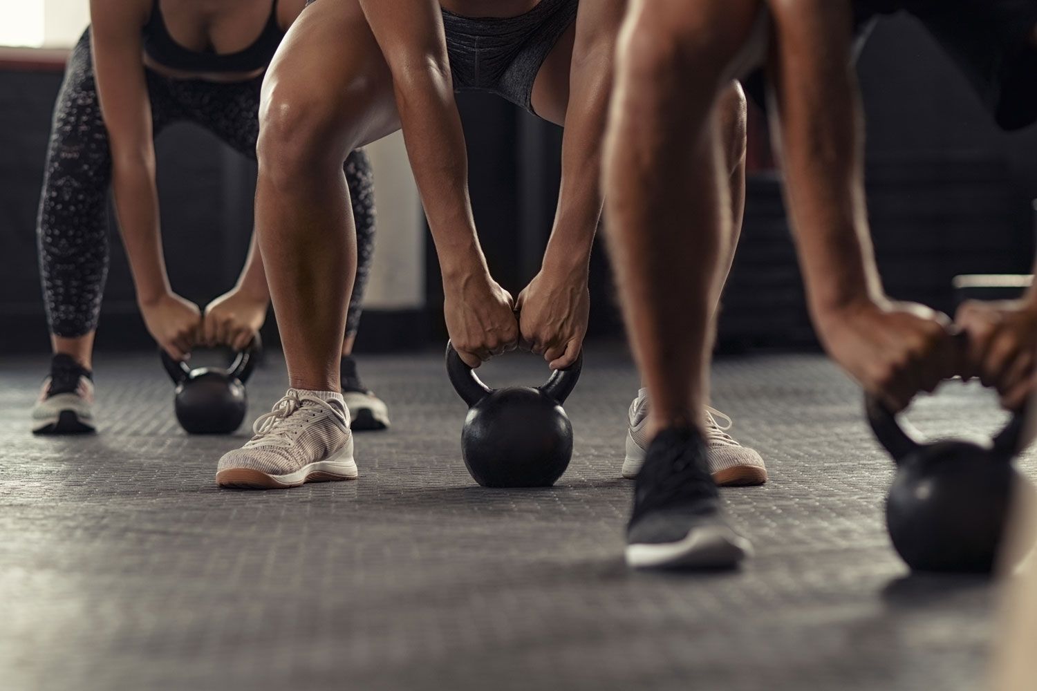 Beine und Hände dreier Menschen, die im Studio nebeneinander Kettlebells vom Boden aufnehmen