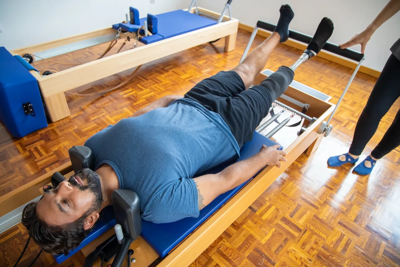 A man lies on a Pilates reformer while a trainer guides the foot bar
