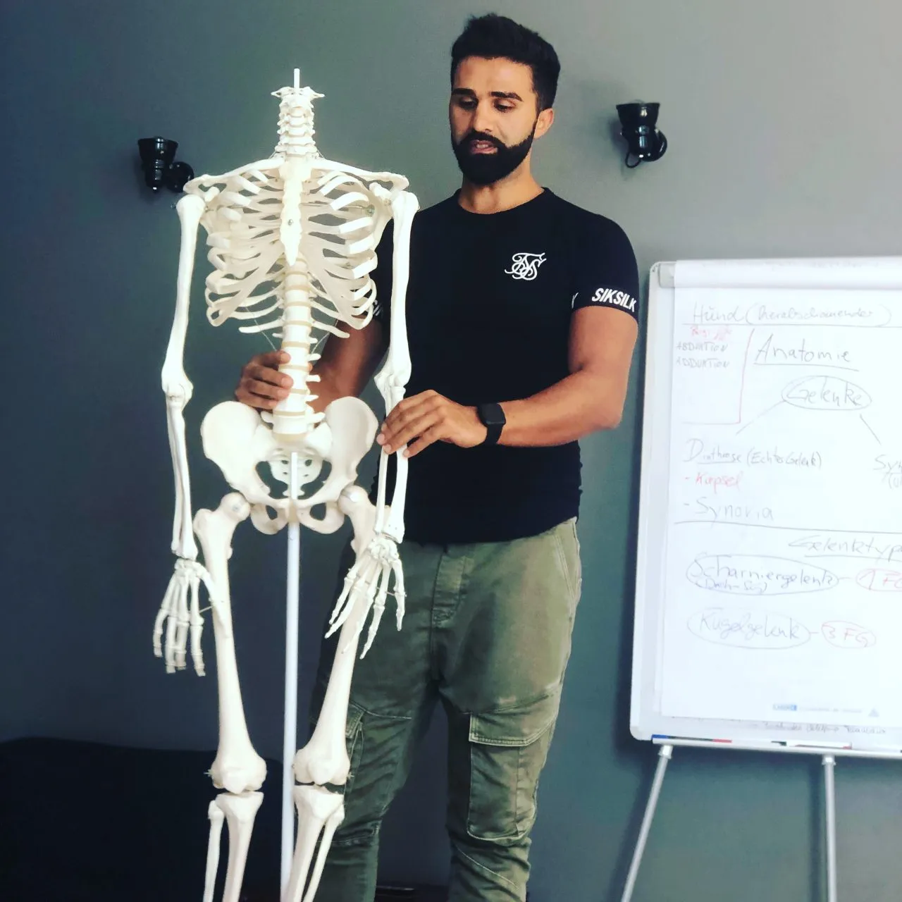 Sohrab Papahn steht neben einem Skelettmodell und zeigt mit der Hand auf die Wirbelsäule, rechts ein Flipchart mit Notizen.